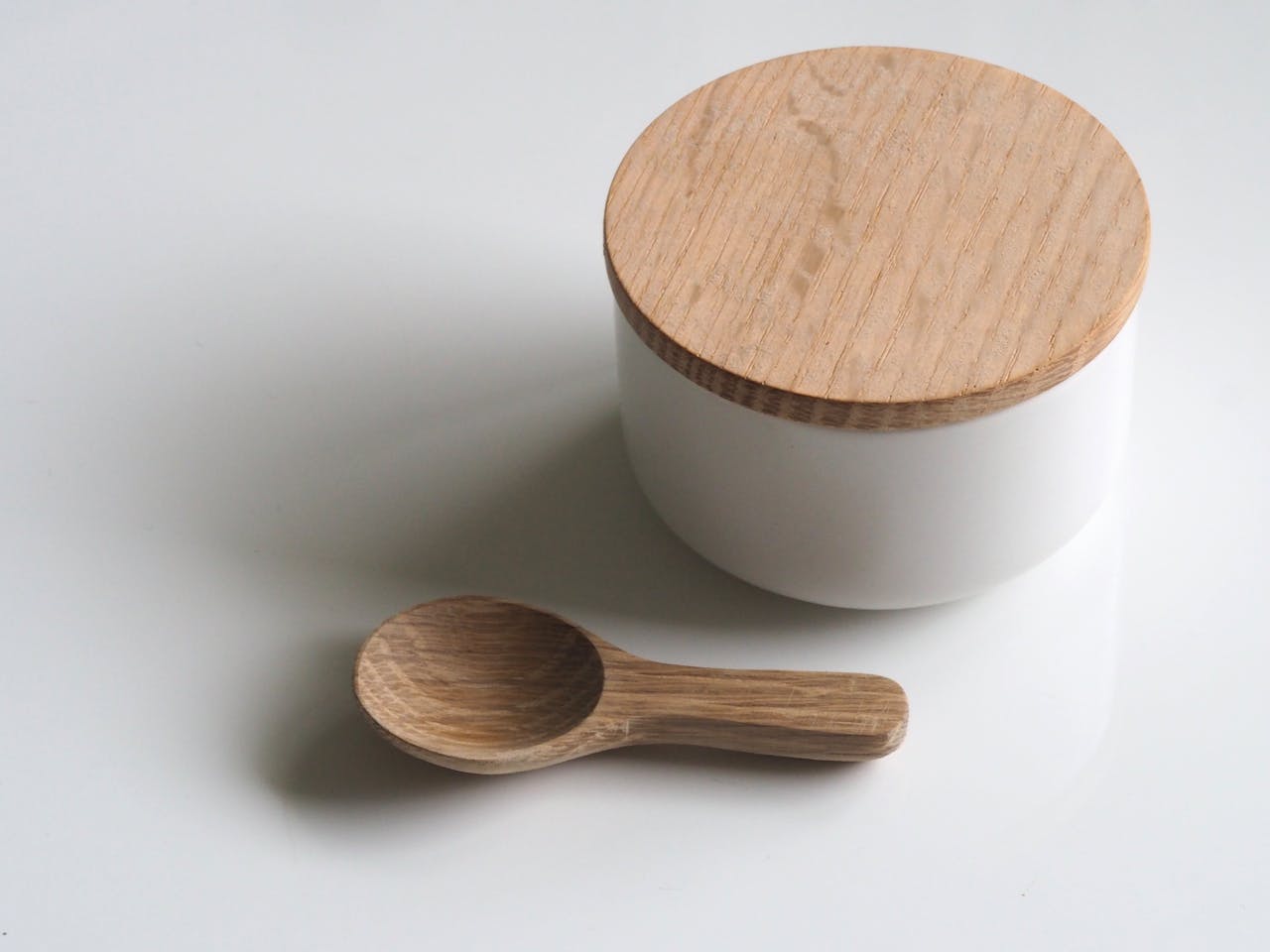 A white round container with a wooden lid and wooden spoon on a white surface. Simple and clean design.